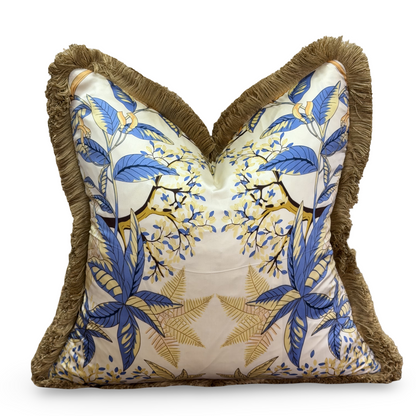 A square decorative throw pillow with a heart-shaped pointed top, featuring a blue and gold tropical floral pattern on a cream background, finished with a brown fringed border.