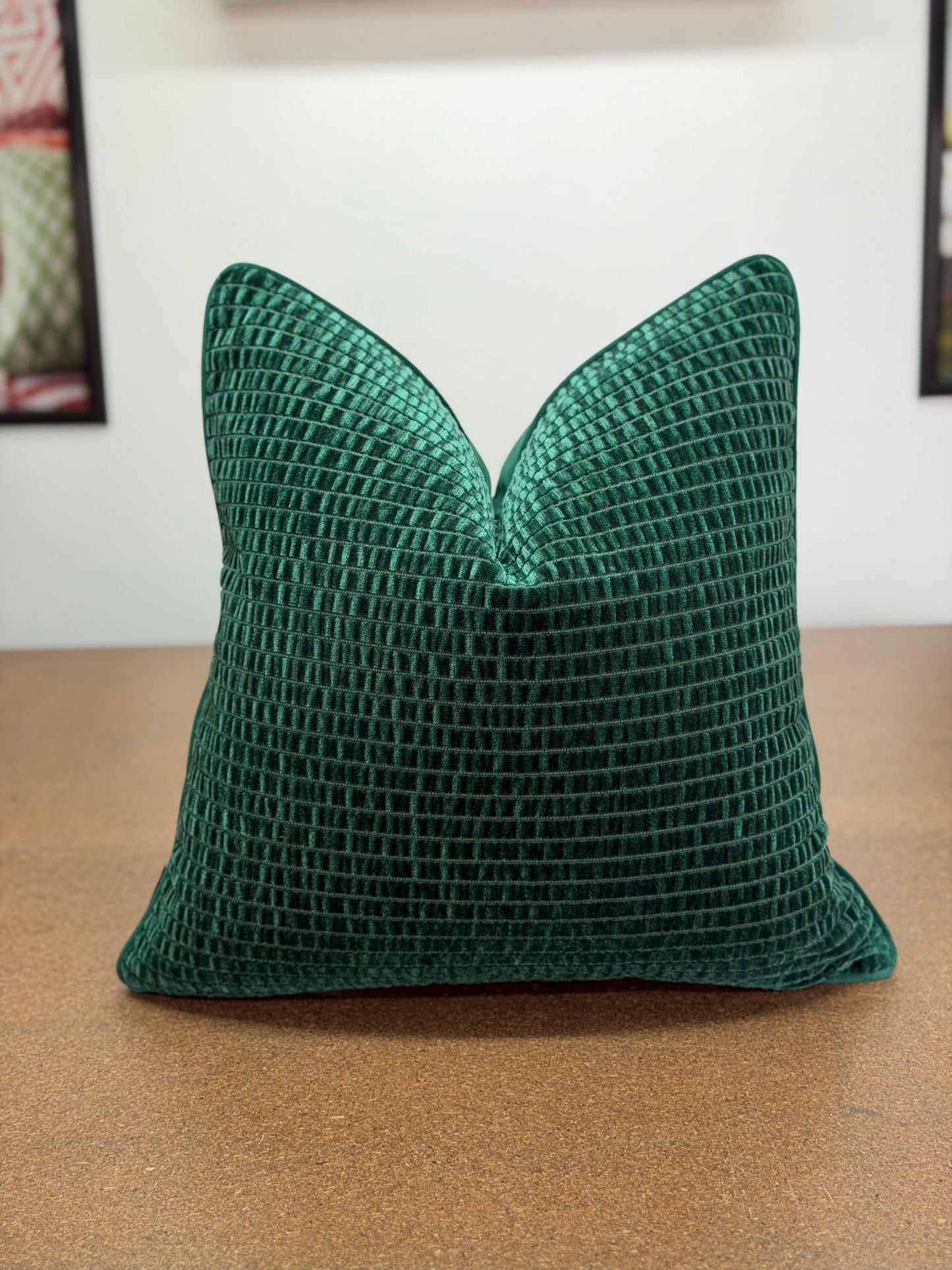 A square emerald green textured throw pillow with a waffle grid pattern, resting on a brown wooden surface against a white wall.