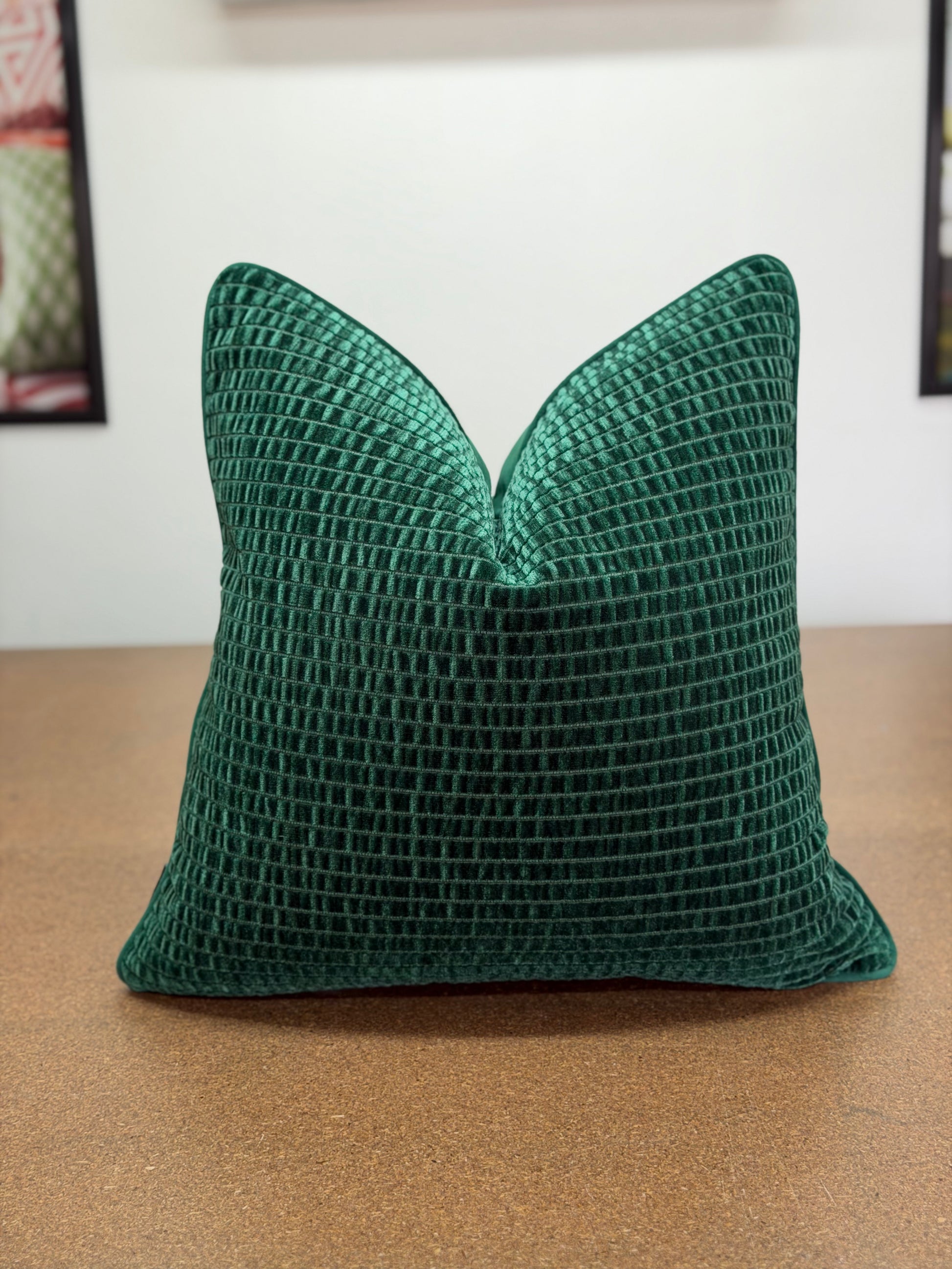 A square emerald green textured throw pillow with a waffle grid pattern, resting on a brown wooden surface against a white wall.