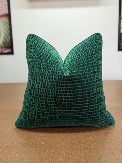A square emerald green textured throw pillow with a waffle grid pattern, resting on a brown wooden surface against a white wall.