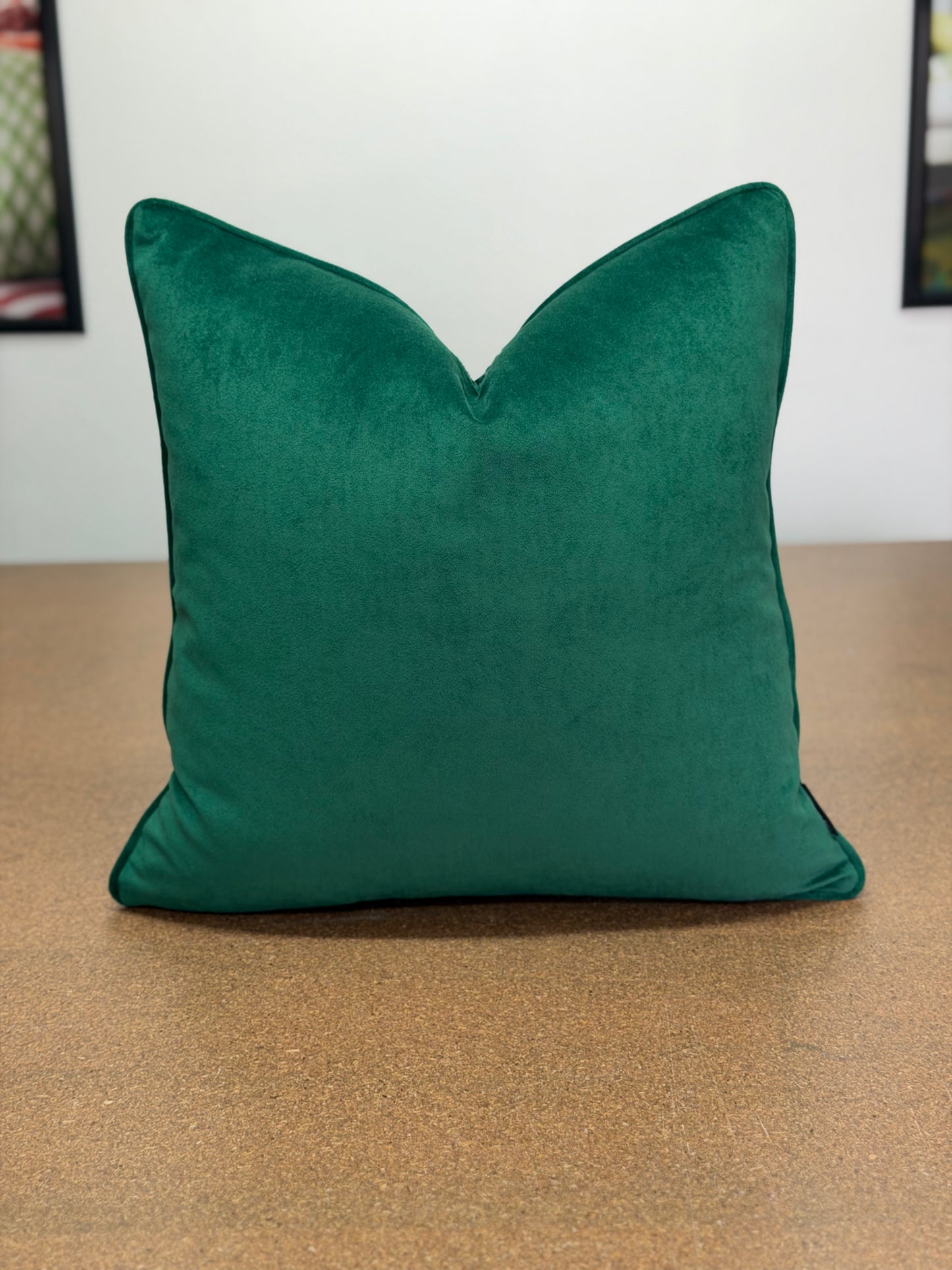 A square emerald green velvet throw pillow with a V-shaped top, resting on a brown textured surface against a white wall.