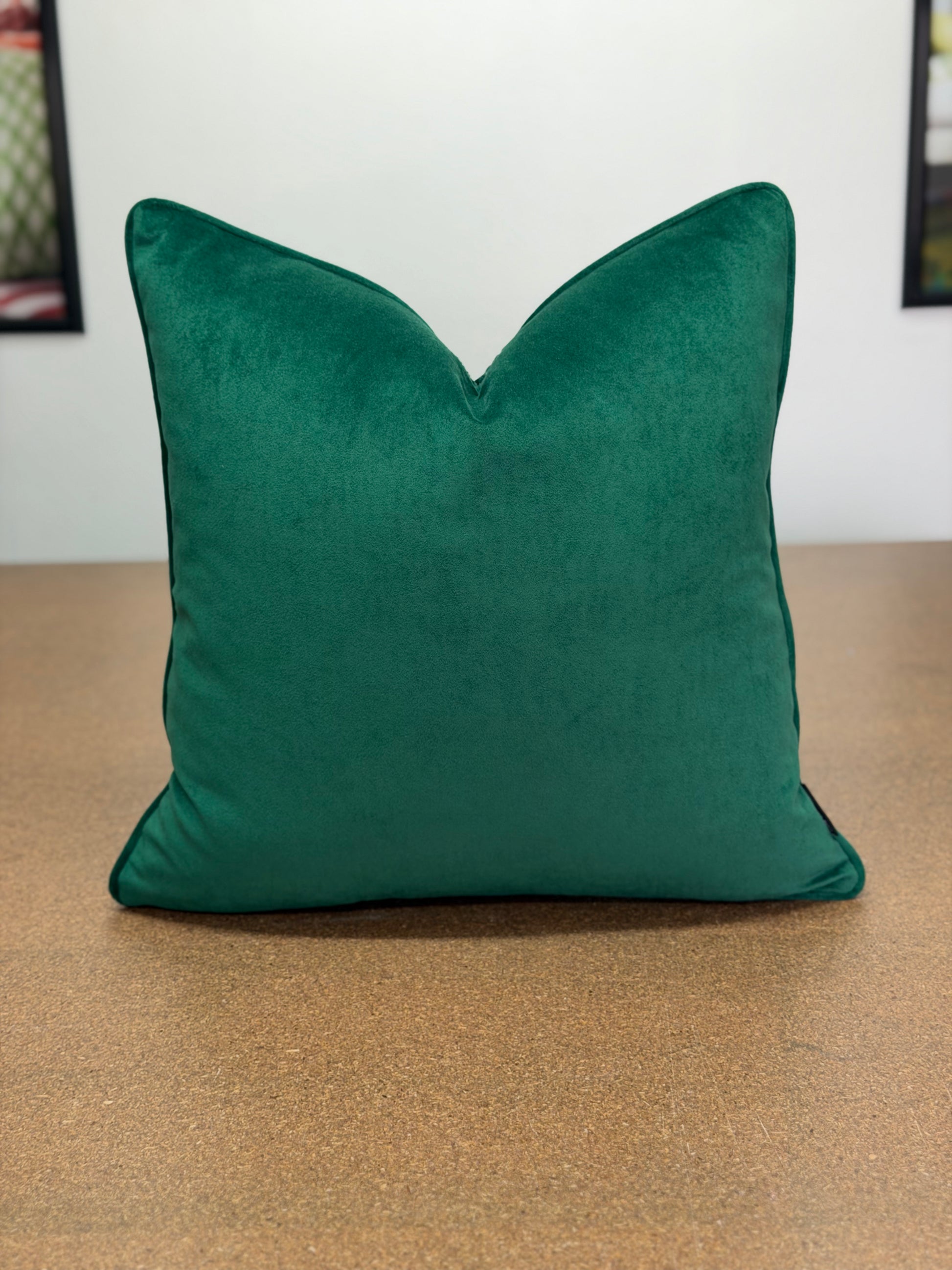 A square emerald green velvet throw pillow with a V-shaped top, resting on a brown textured surface against a white wall.