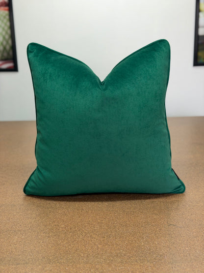 A square emerald green velvet throw pillow with a V-shaped top, resting on a brown textured surface against a white wall.