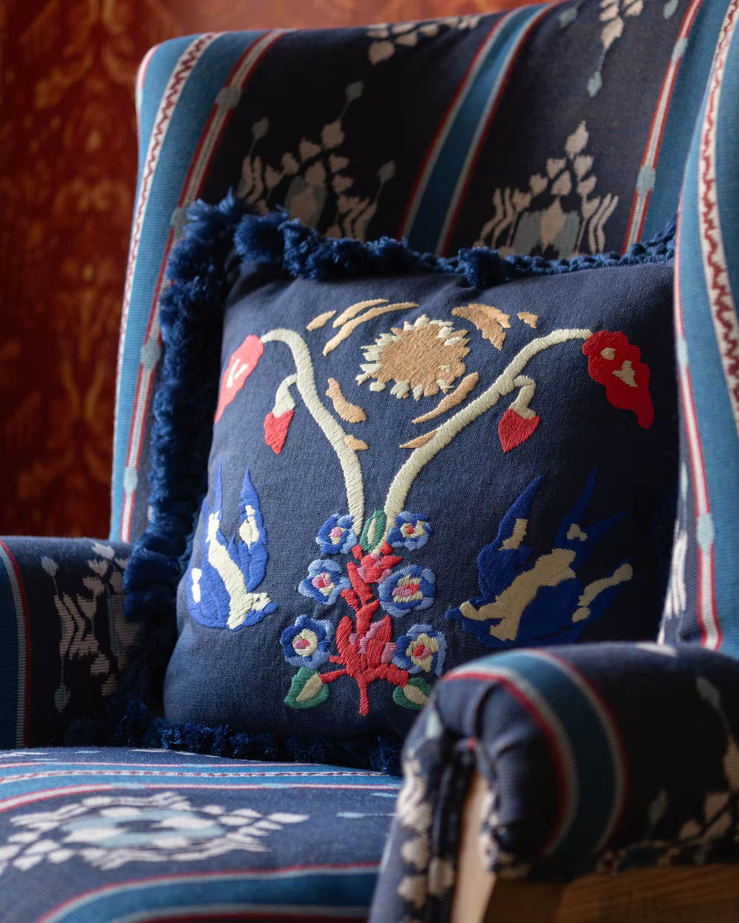 A navy blue embroidered throw pillow with colorful floral and bird motifs, resting on a matching patterned upholstered armchair.