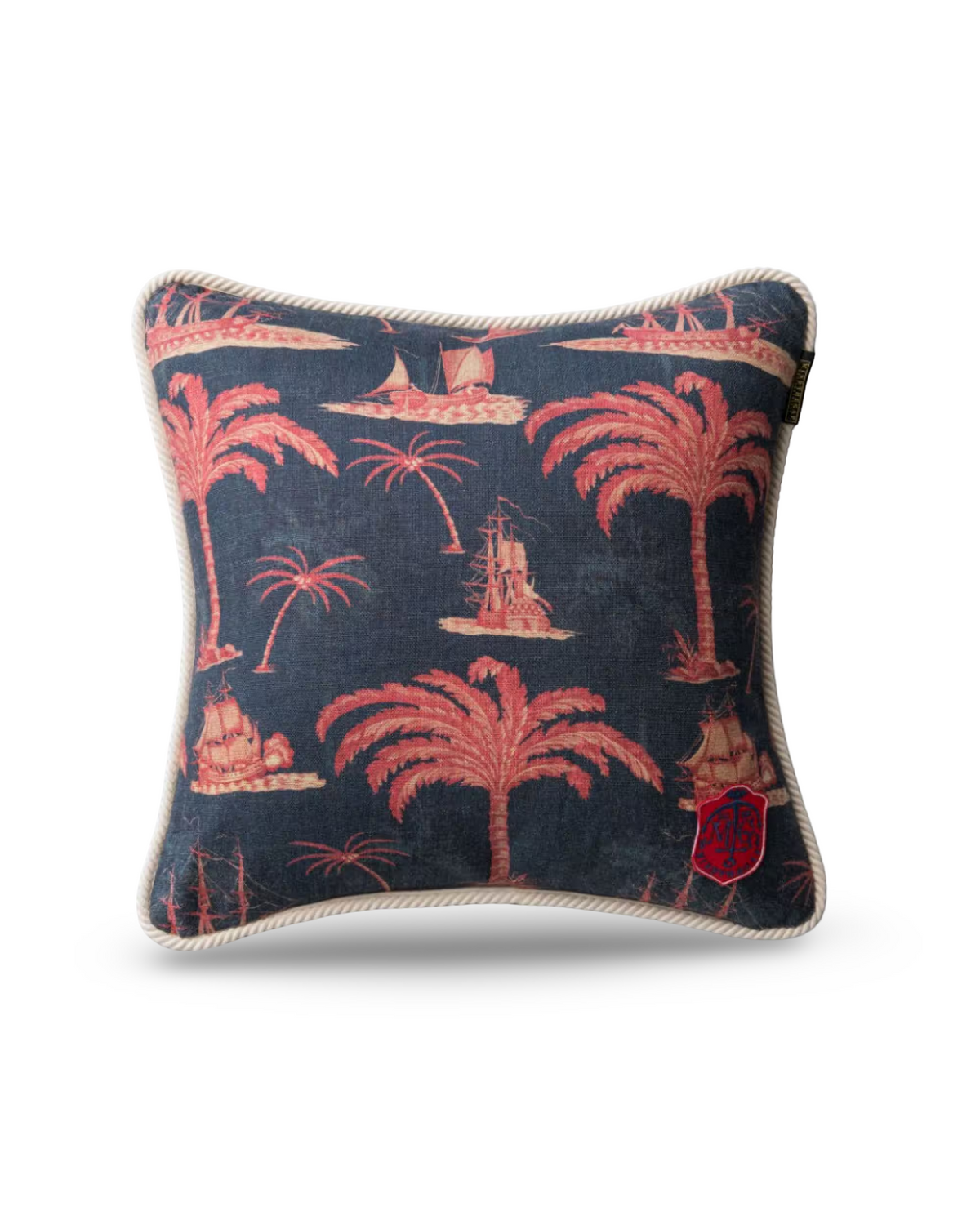 A square decorative throw pillow featuring a navy blue and pink tropical toile pattern with palm trees, ships, and a shield emblem, finished with a cream-colored twisted rope trim.