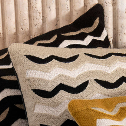 A close-up of a textured throw pillow with a black and cream chevron pattern, layered over another pillow with a black and cream abstract pattern, with a mustard yellow accent pillow in the foreground, set against a light wood slatted background.