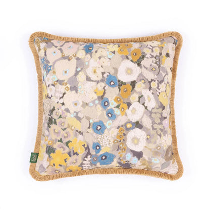 A square decorative throw pillow with a beige background and a dense, multicolored floral pattern in blue, yellow, white, and gray. The pillow features a natural jute fringed border and a small green brand tag on the bottom left corner, photographed against a seamless white background.